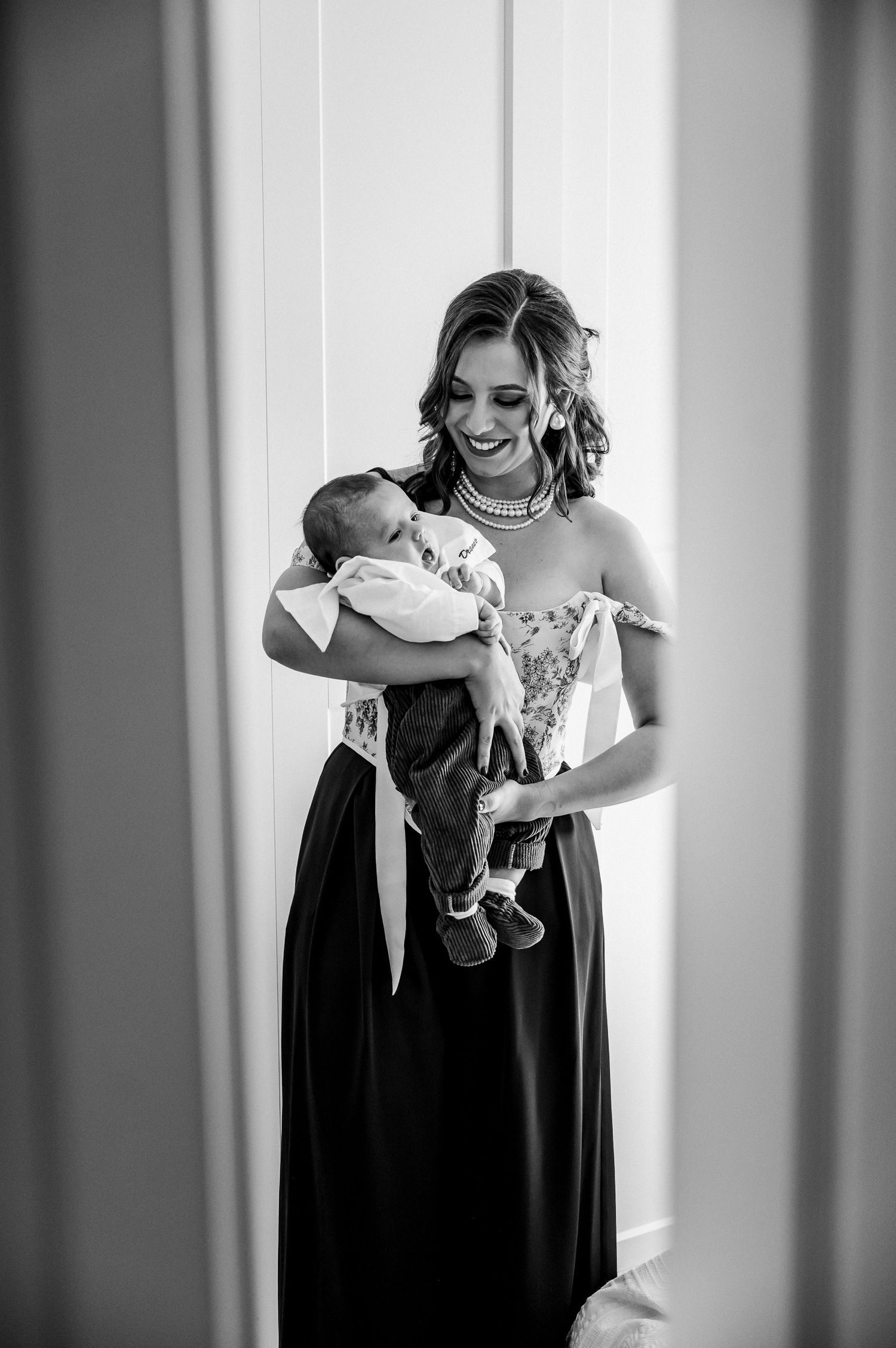 Smiling bridesmaid in black gown holds newborn baby in black and white wedding photo.