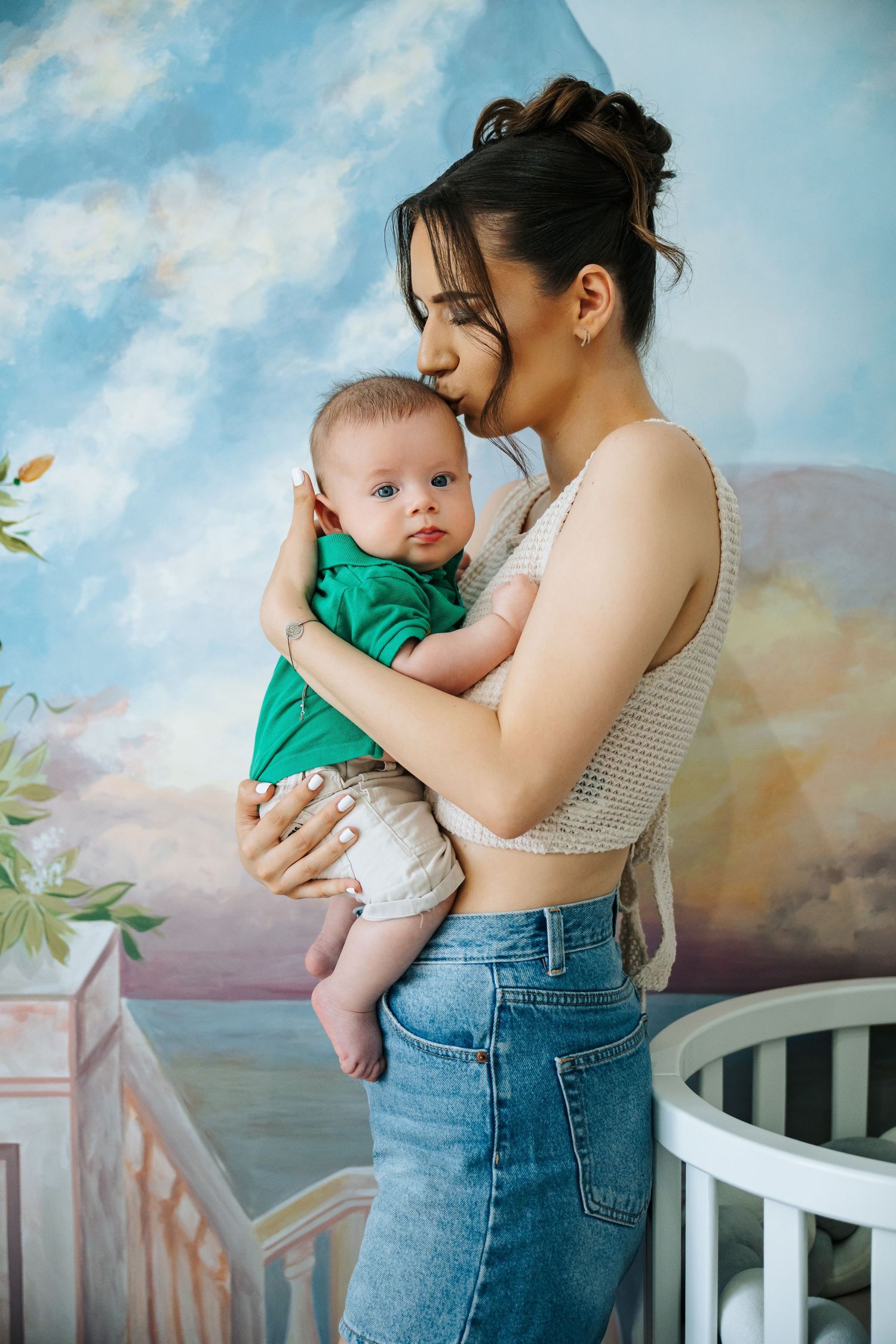 Young brunette mother in green top and jeans lovingly holds her baby in a decorated nursery room.