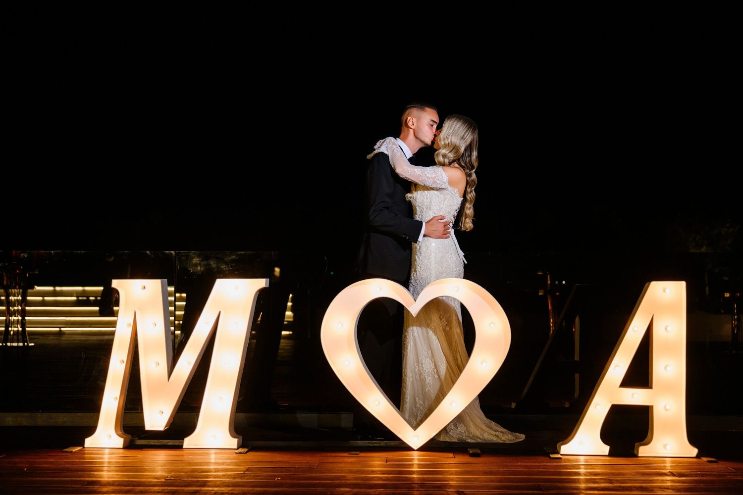 Couple shares tender embrace between glowing marquee letters M and A with heart symbol at night wedding.