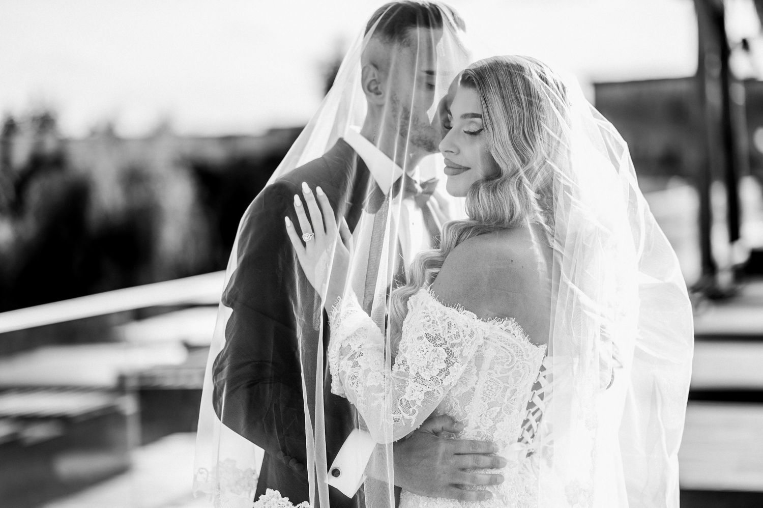 Black and white romantic photo of bride and groom embracing under flowing wedding veil.