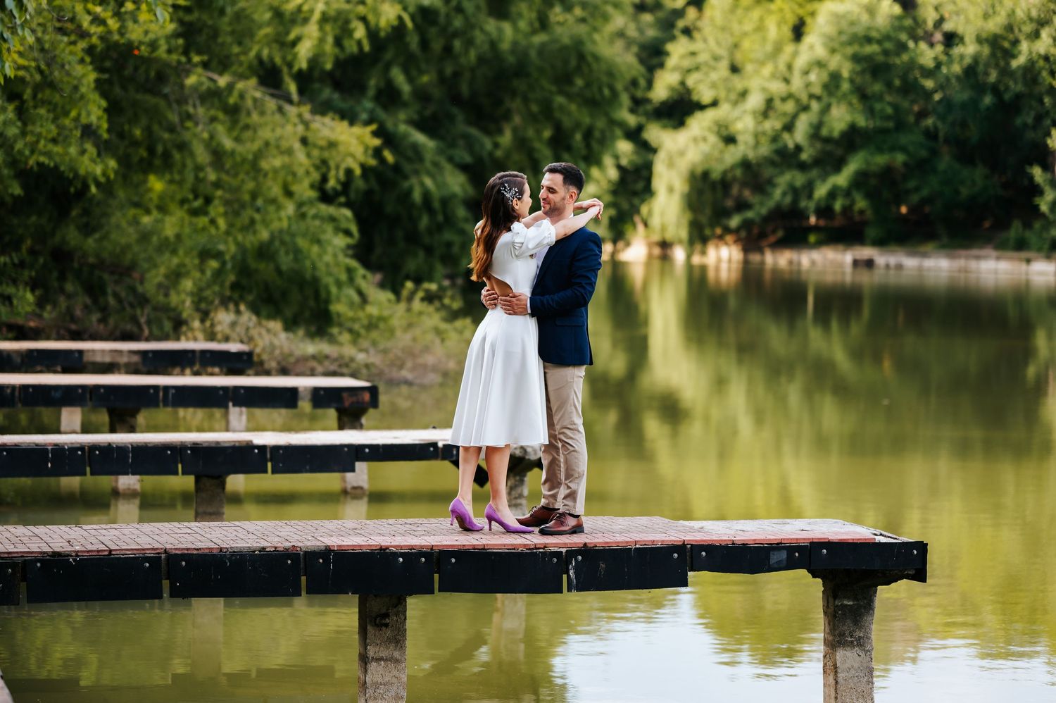 Couple embracing on a wooden dock surrounded by a serene green lake and lush trees.
