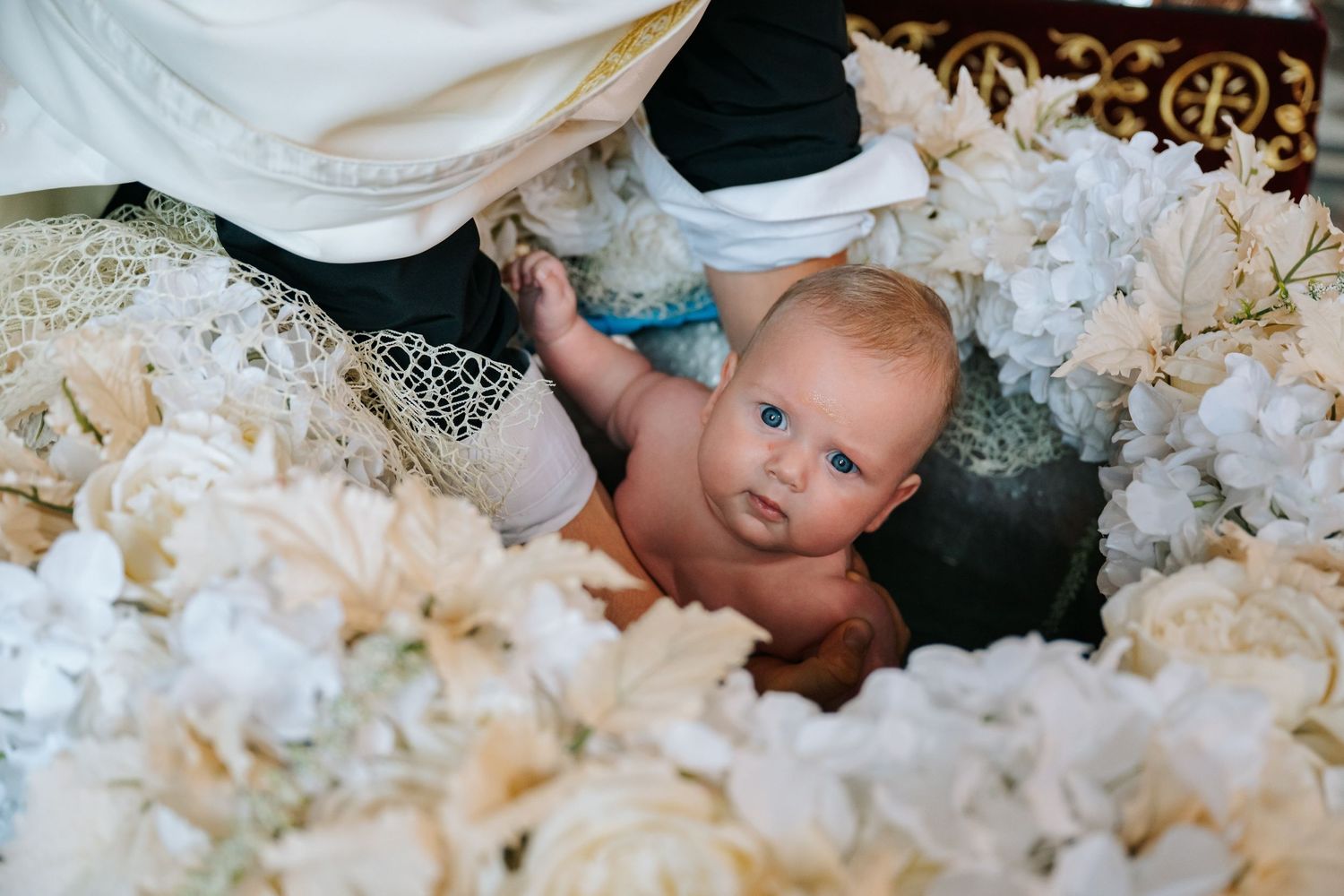 Baby being baptized in a flower-adorned baptismal font during a religious ceremony.