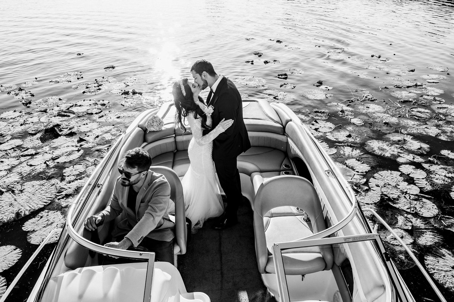 Bride and groom share a kiss on a motorboat surrounded by lily pads in a black and white aerial photo.