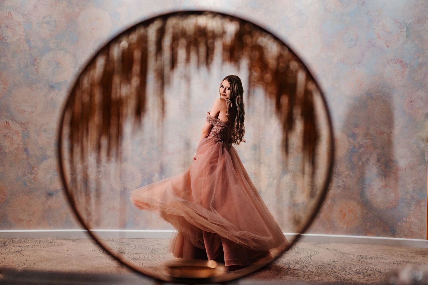 Woman in flowing pink tulle gown twirling inside a circular frame against a rustic textured backdrop.