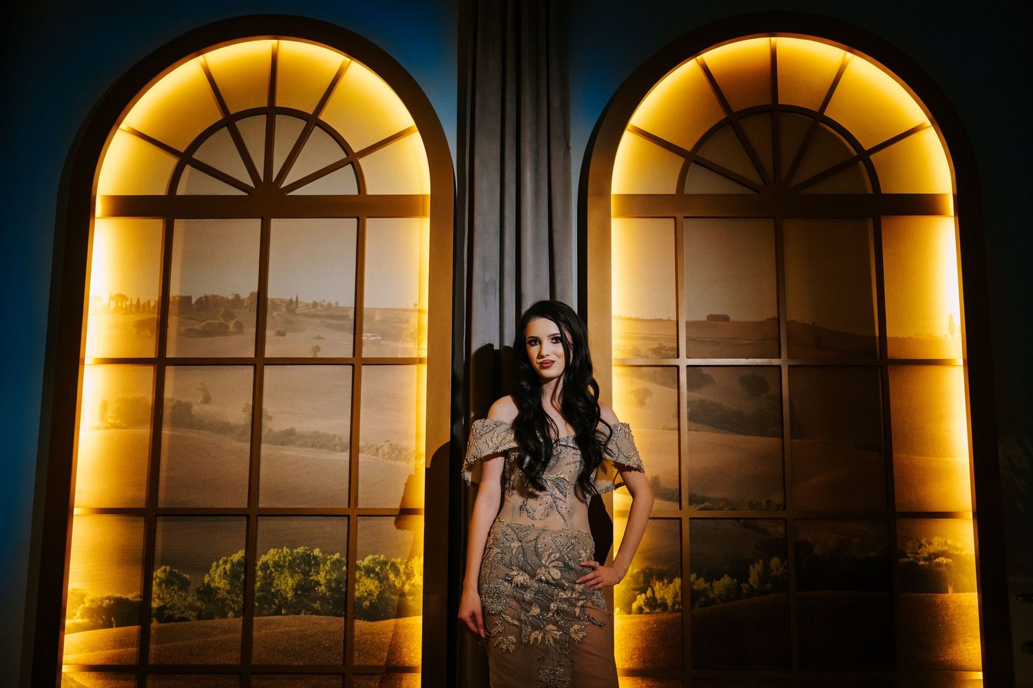Woman in elegant gown posing between two arched golden-lit windows with vineyard backdrop.