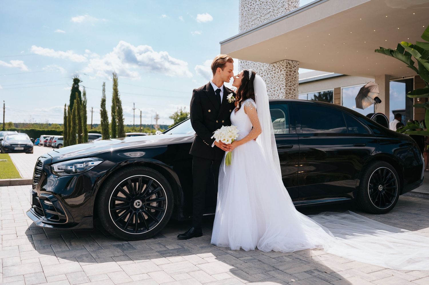 Bride and groom kissing beside a black Mercedes-Benz luxury car on their wedding day outdoors.
