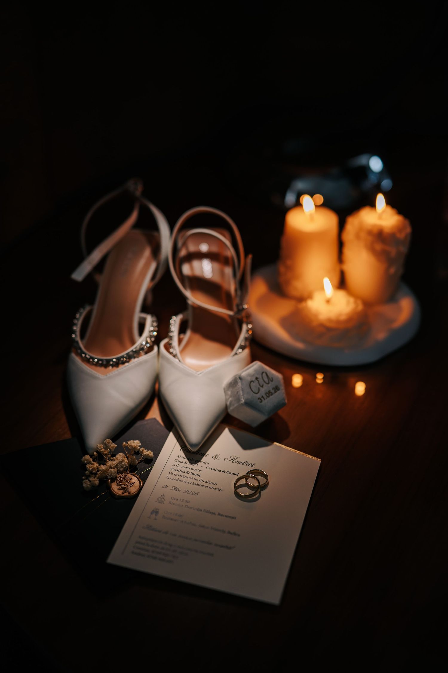 Wedding flat lay featuring white bridal heels, gold rings, invitation card, and glowing candles in dark setting.