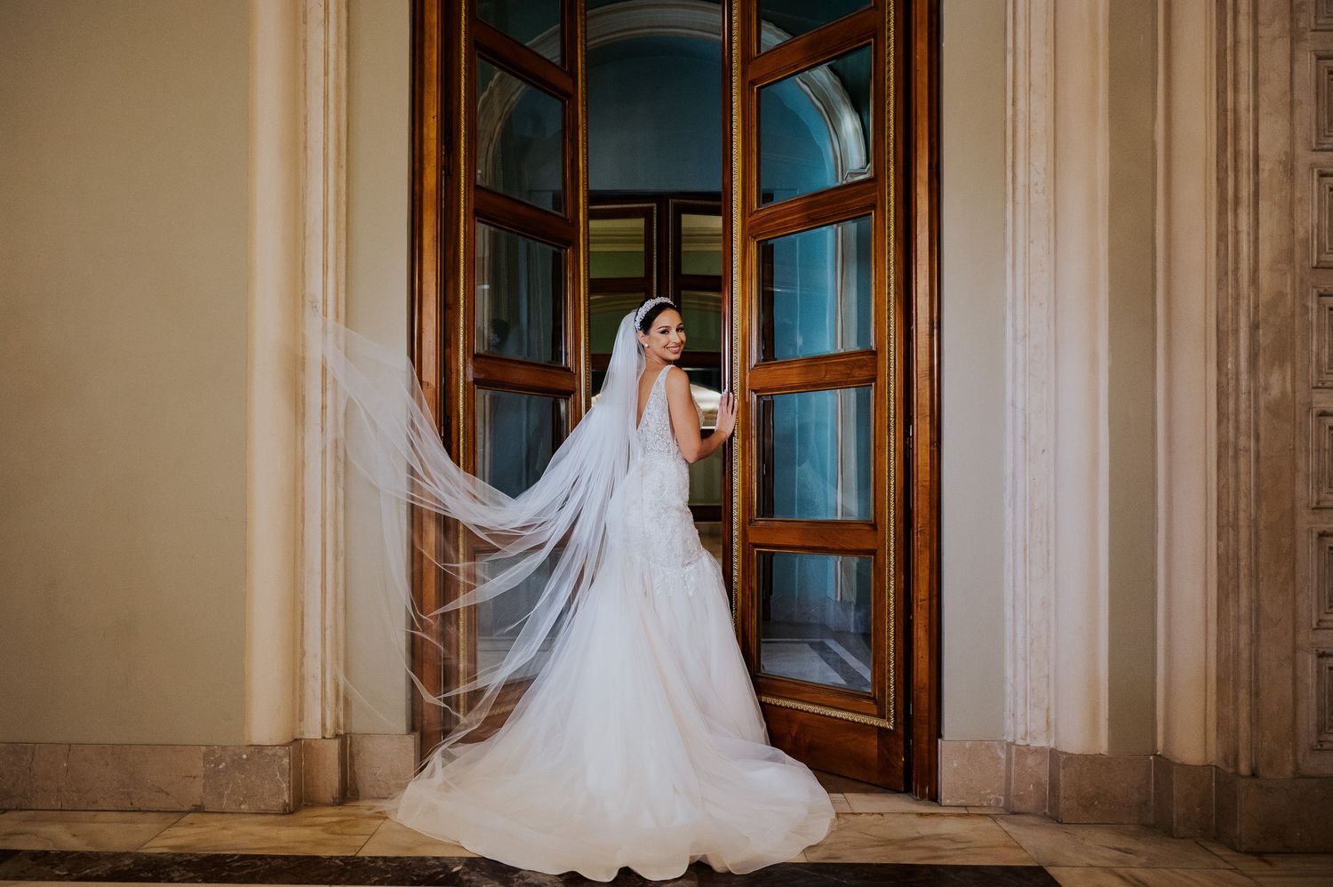 Bride in white wedding gown and flowing veil stands before ornate wooden doors at elegant venue.