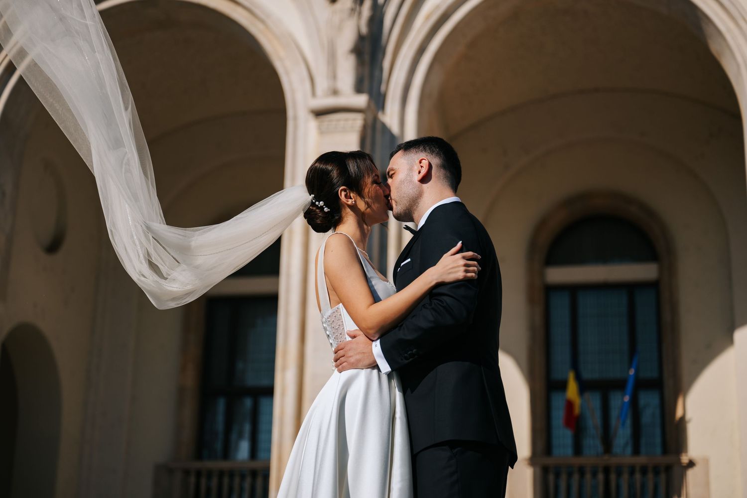 Bride and groom share a romantic kiss outdoors, with veil flowing in the wind against elegant arched architecture.