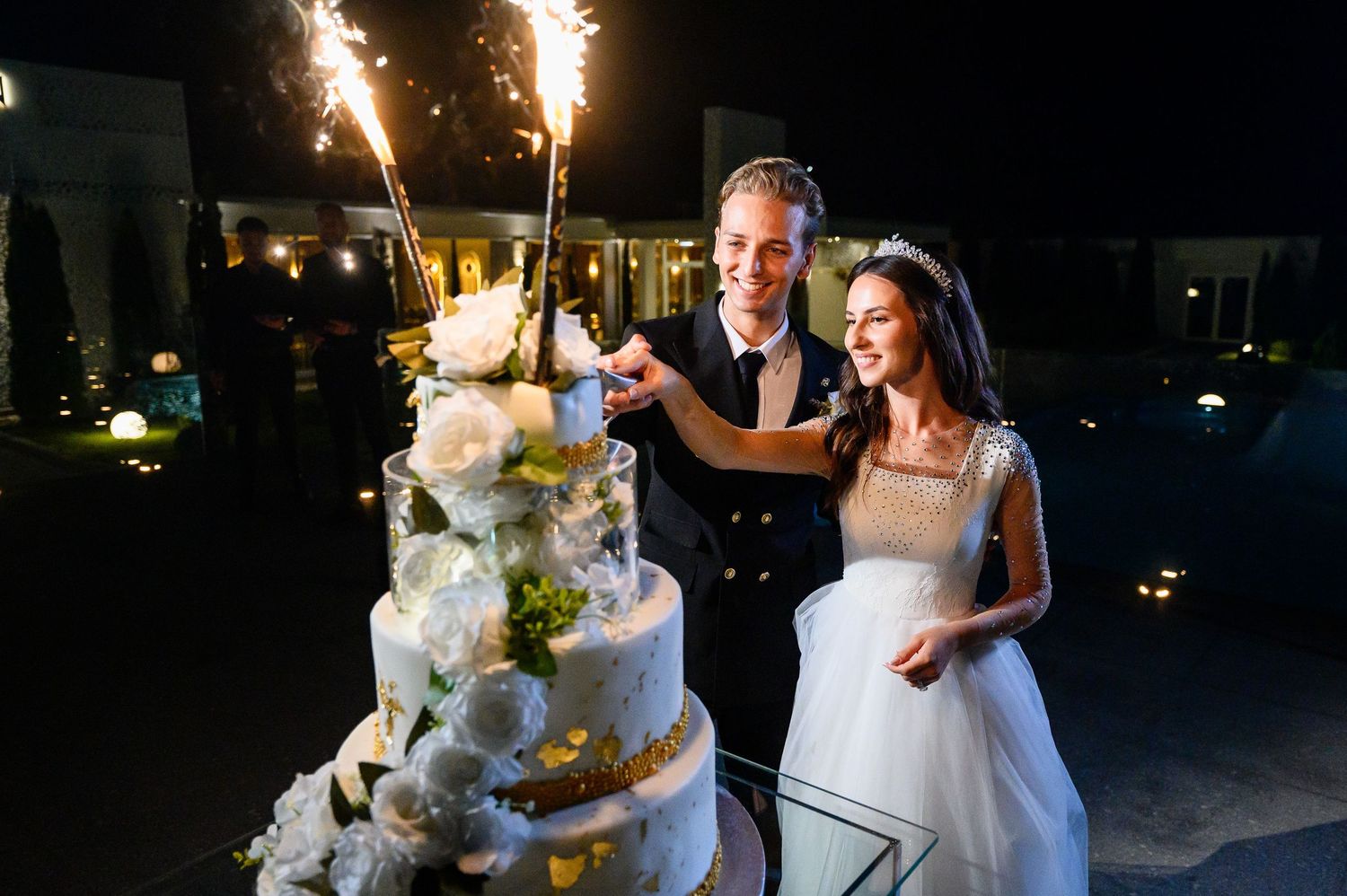 Bride and groom cutting a white floral wedding cake with sparklers at an outdoor night reception.