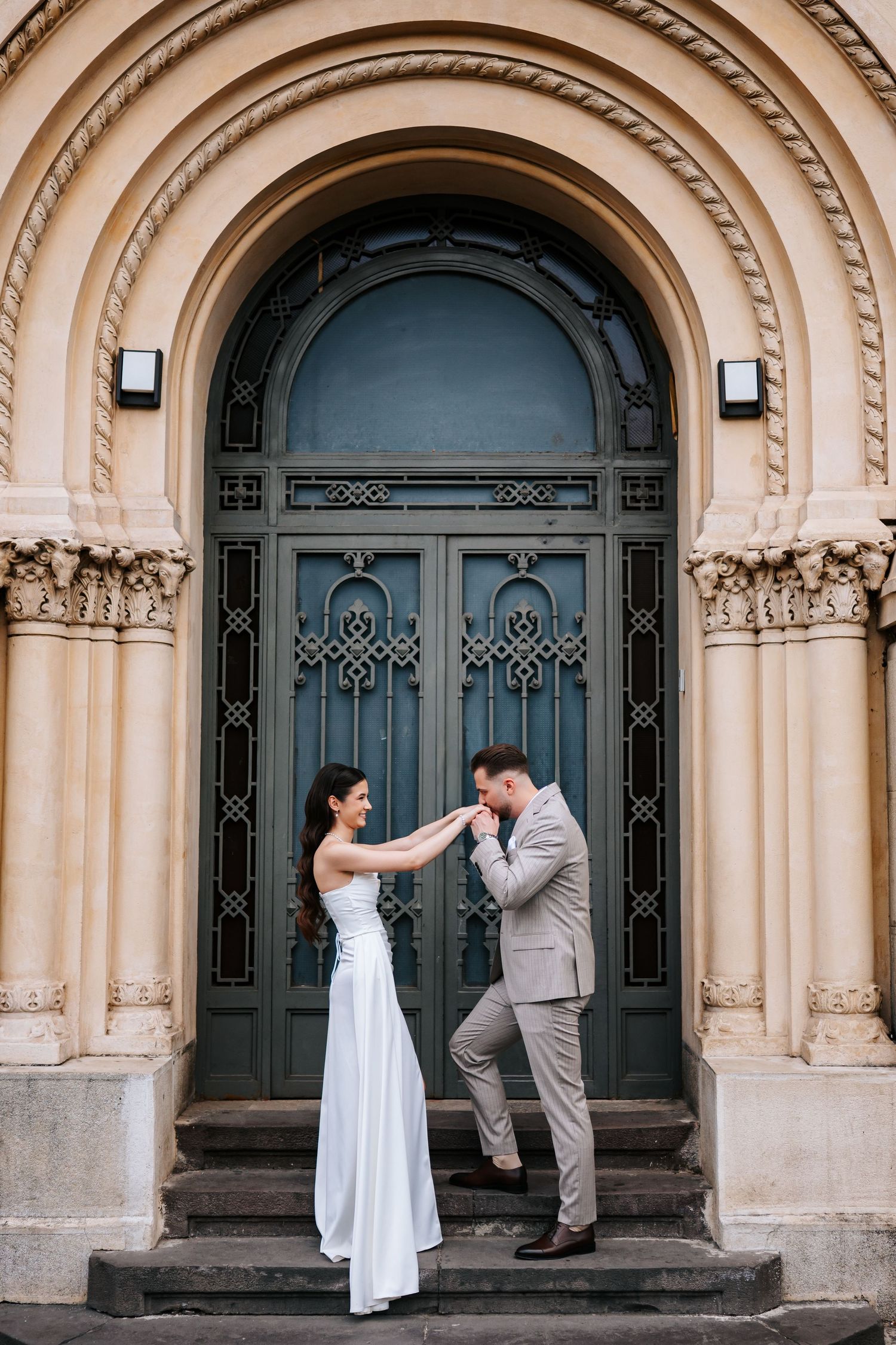 Bride and groom stand on steps before ornate arched green door as groom kisses her hand.