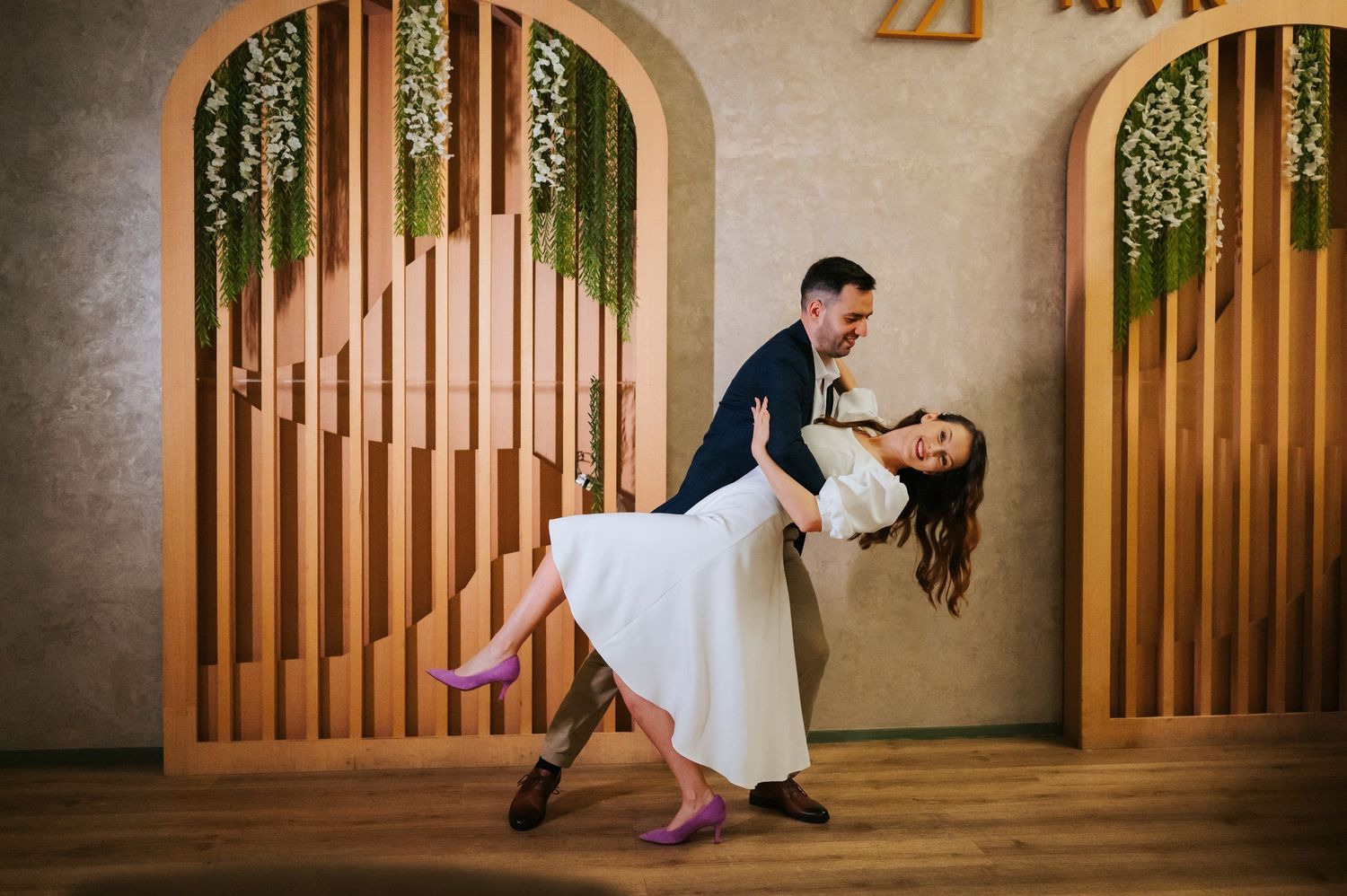 Couple performing elegant wedding dance dip in front of decorative wooden arch with hanging greenery.