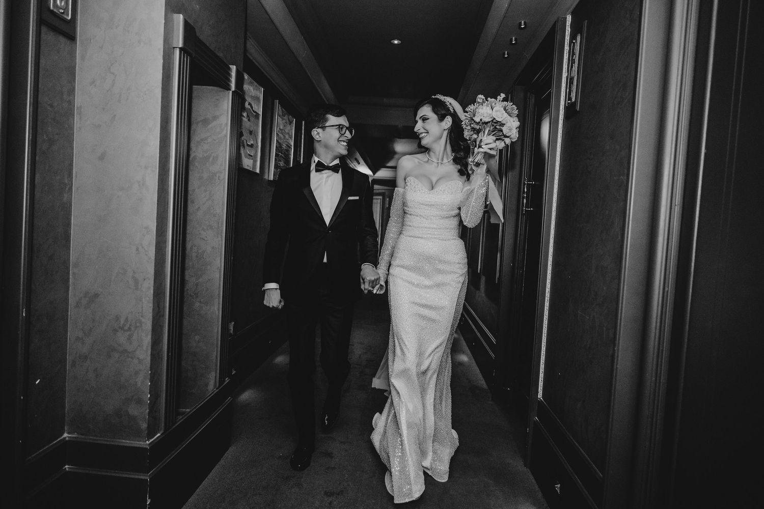 Bride and groom walk hand in hand down elegant hotel hallway in classic black and white wedding portrait.