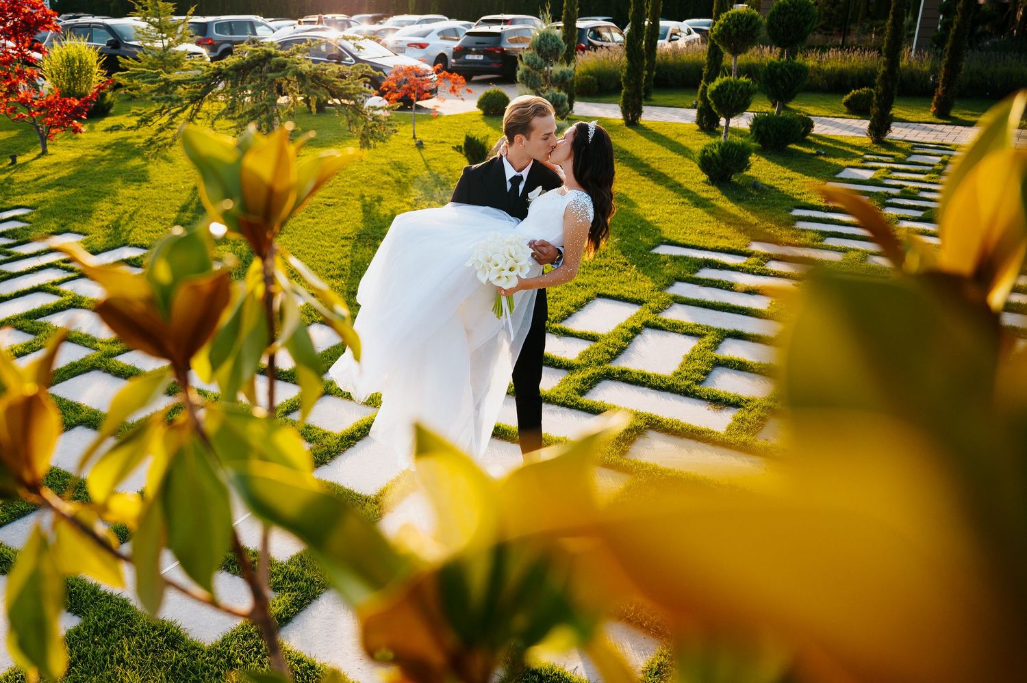 A groom in a black suit lifts his bride in a white wedding dress outdoors on a garden path surrounded by lush greenery.