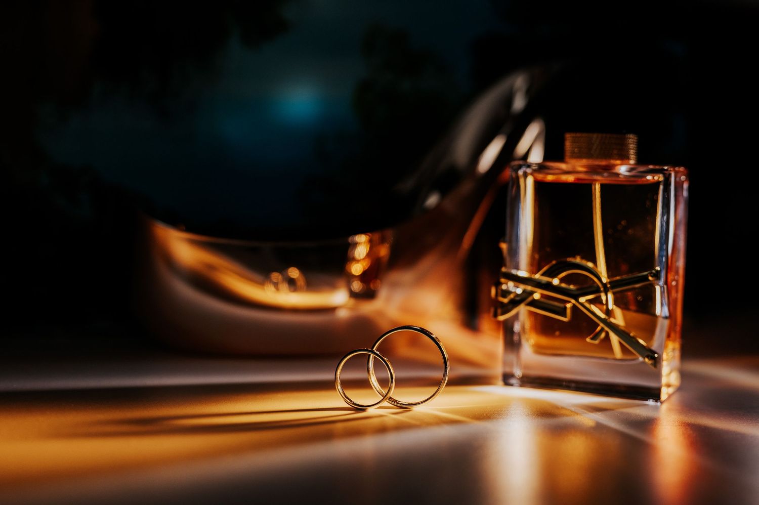 Gold wedding rings beside a perfume bottle and high heel shoe in dramatic warm lighting.