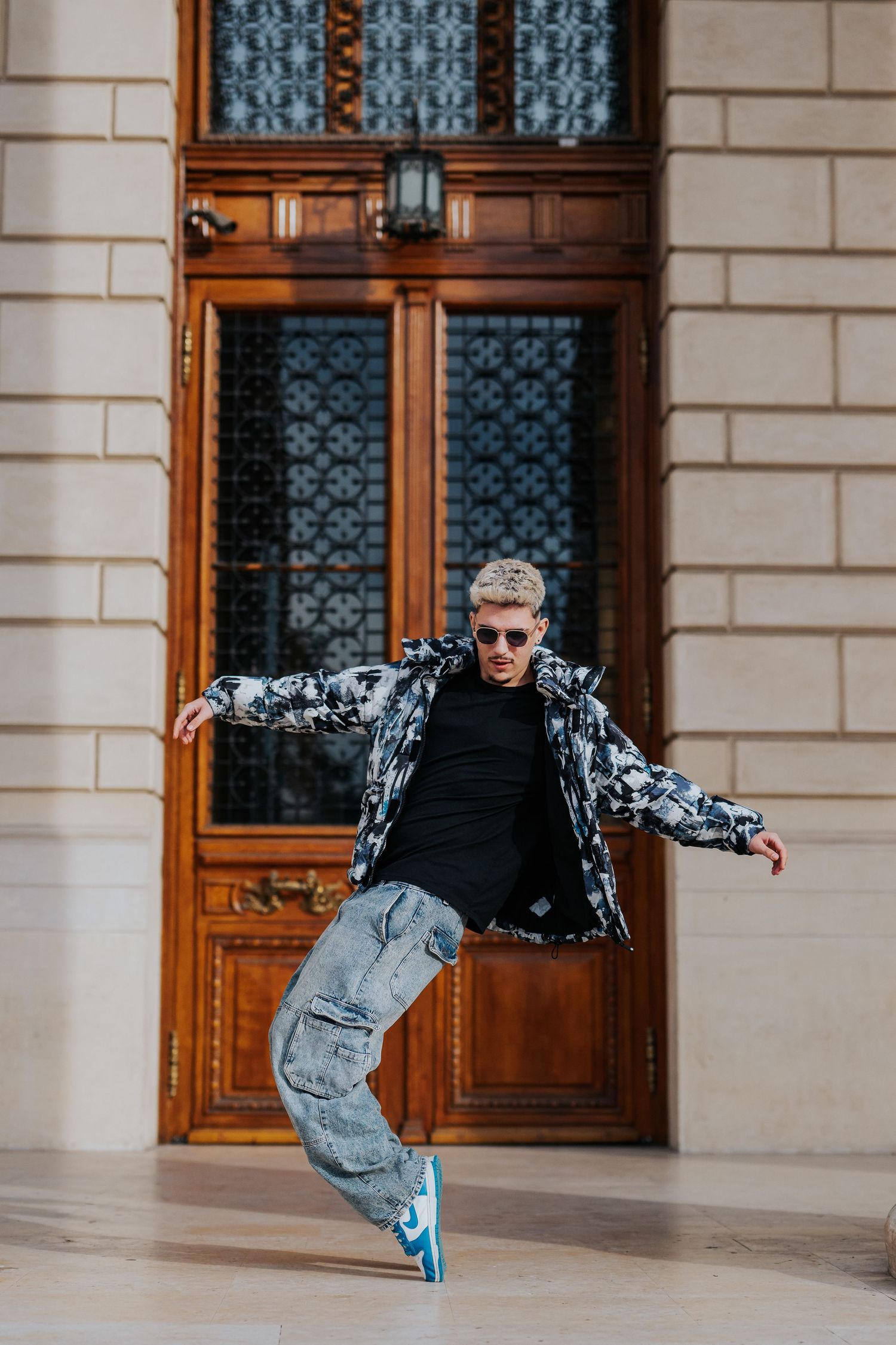 Young man in patterned jacket, cargo jeans, and blue sneakers striking a dance pose before ornate wooden doors.