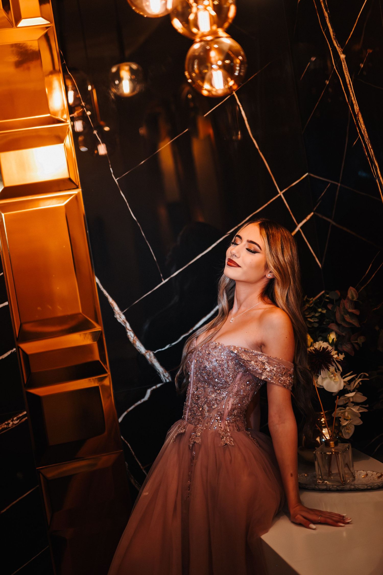 Elegant woman in a beaded off-shoulder mauve ball gown poses in a luxurious dark marble interior with gold accents.