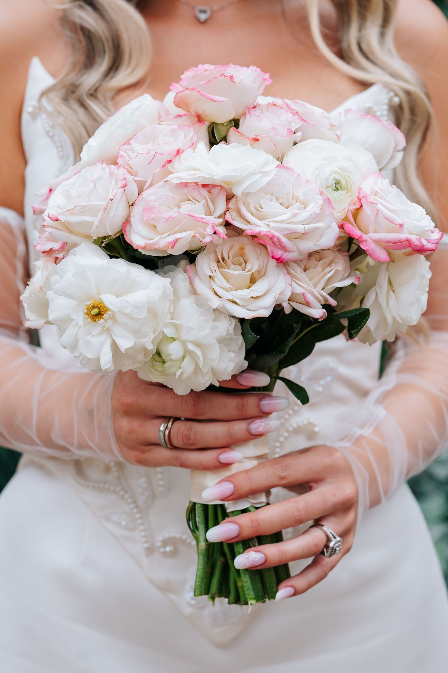 Bride holds elegant bouquet of white and pink roses with manicured hands wearing wedding and engagement rings.