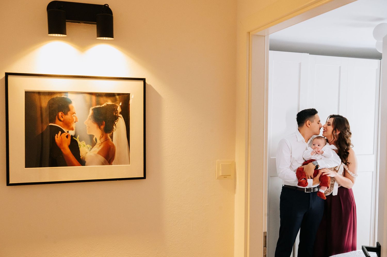 Happy couple kisses while holding baby in doorway, with their framed wedding portrait displayed on wall.