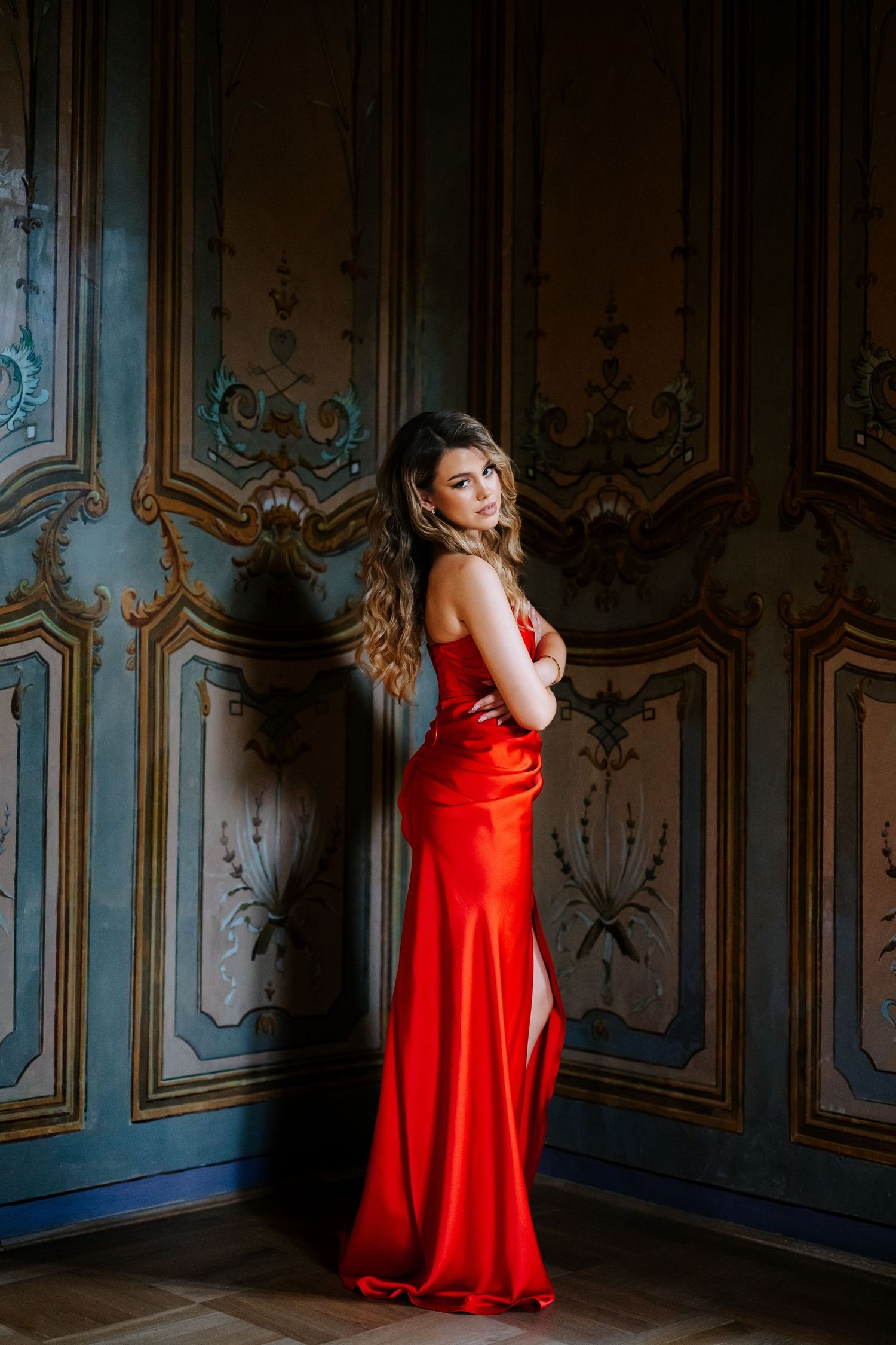 Blonde woman in red satin gown posing against ornate vintage painted wall panels.