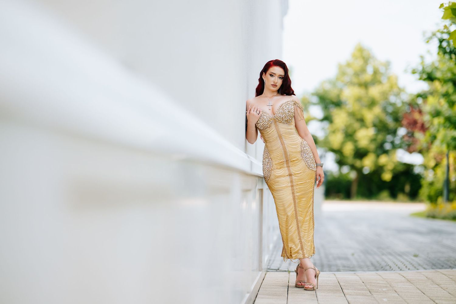 Woman in elegant gold midi dress posing gracefully outdoors near white wall with green trees in background.