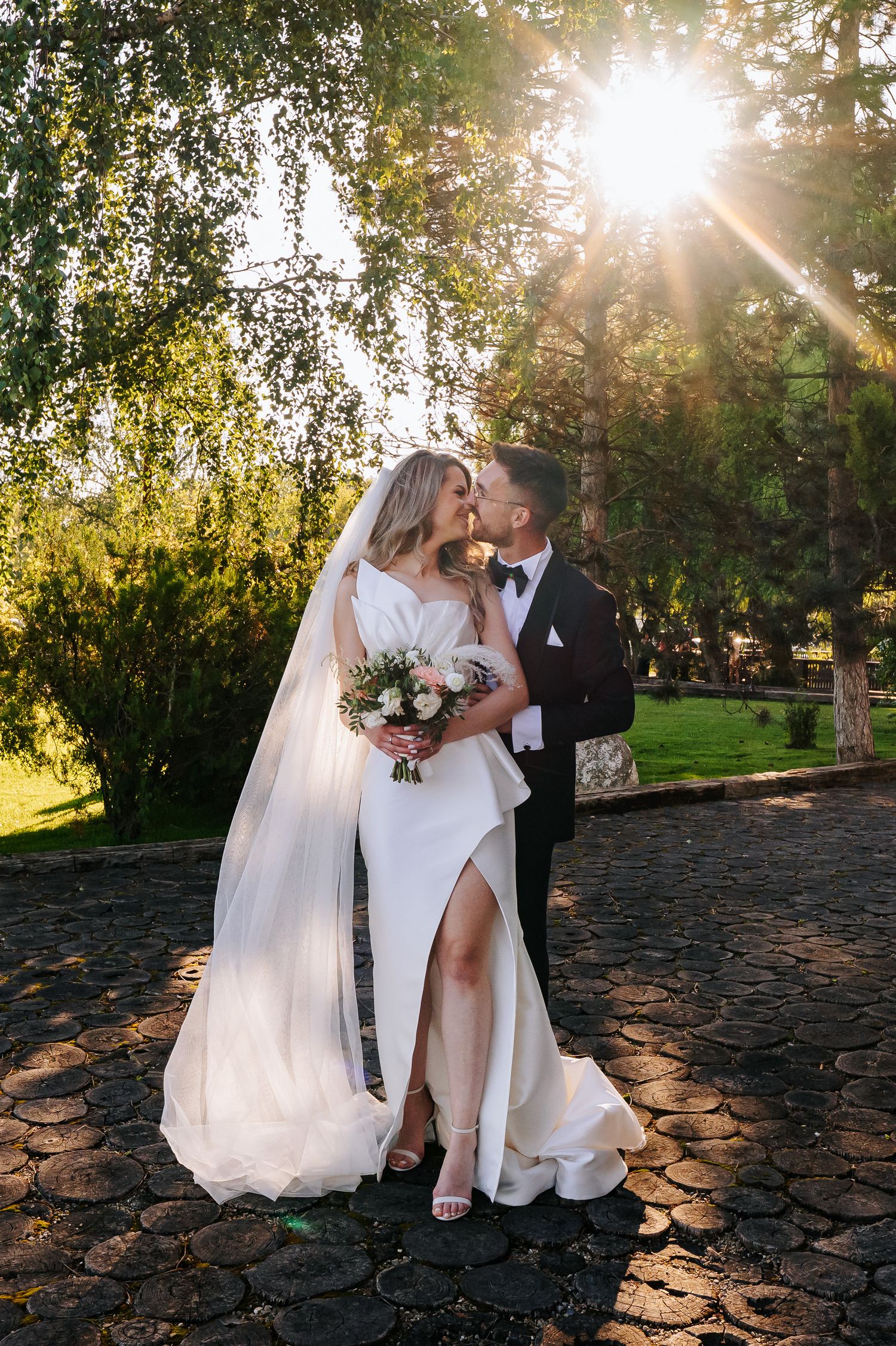 Bride and groom share a romantic moment outdoors, with sunlight streaming through trees, bride holding a bouquet.