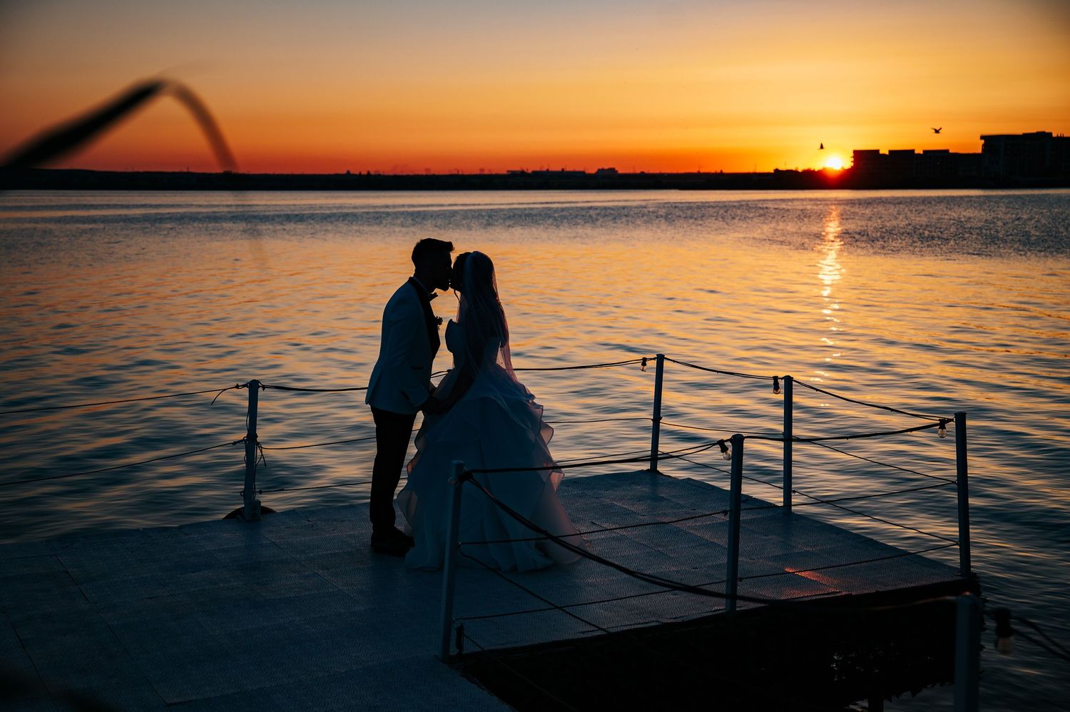 Couple sharing a romantic kiss on a dock at sunset over calm water, silhouetted against an orange sky.
