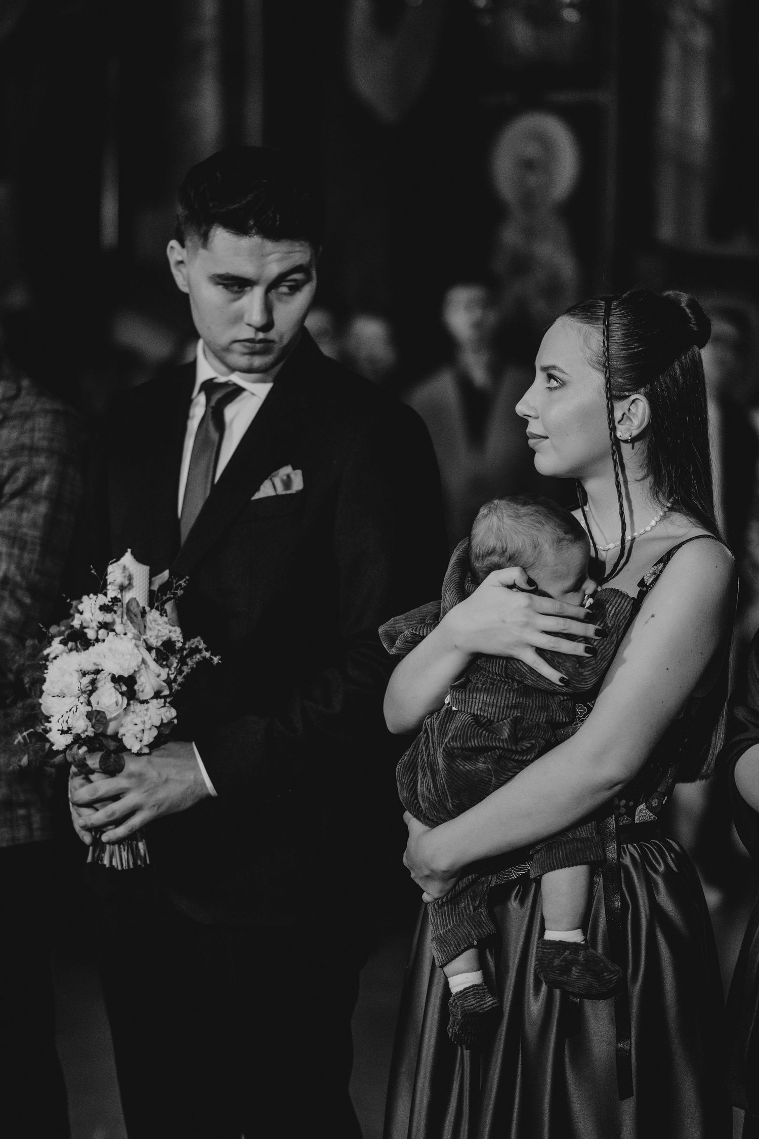 Black and white photo of a young couple in formal attire holding a baby during a church ceremony.