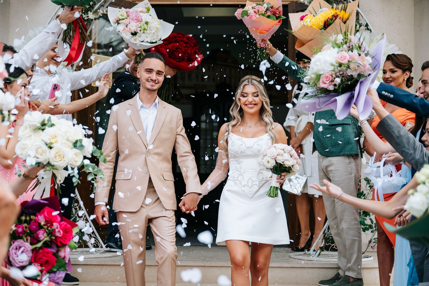 Newlyweds exit ceremony hall holding hands as guests celebrate with confetti and colorful bouquets.