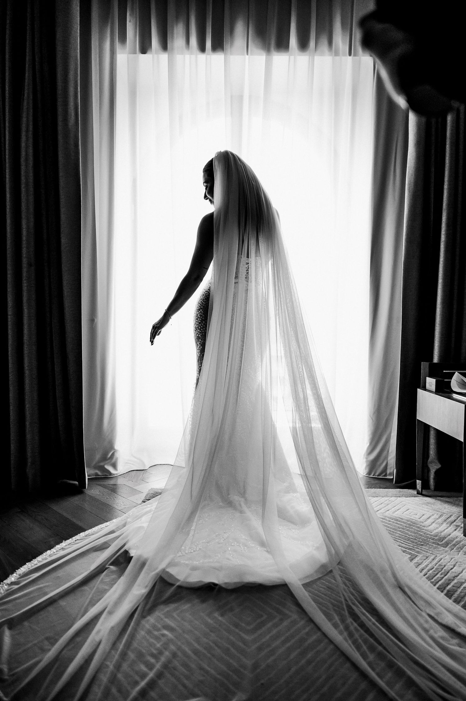Dramatic black and white silhouette of bride in long veil standing before bright window in wedding gown.