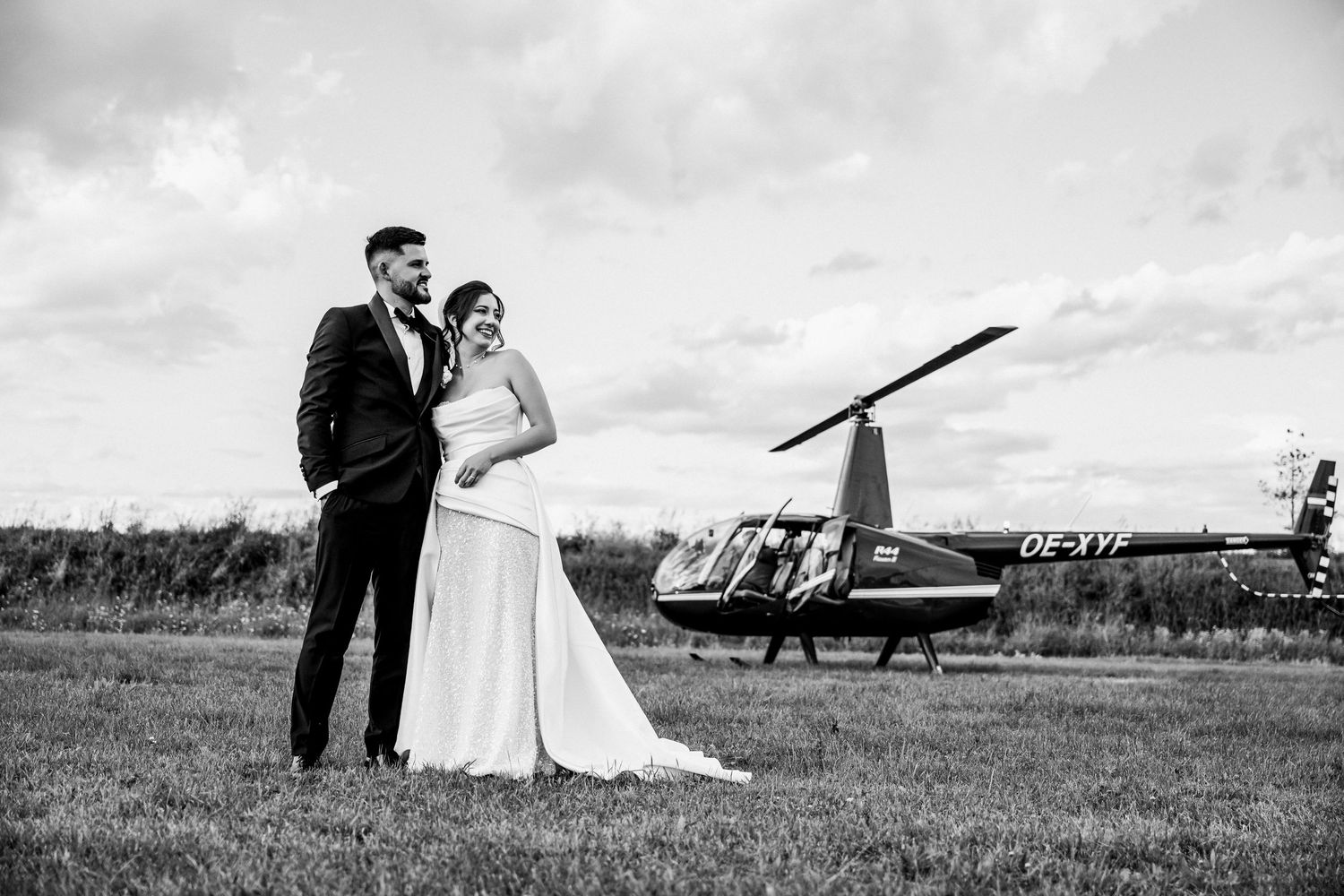 Black and white wedding photo of bride and groom posing in a field with a helicopter in the background.