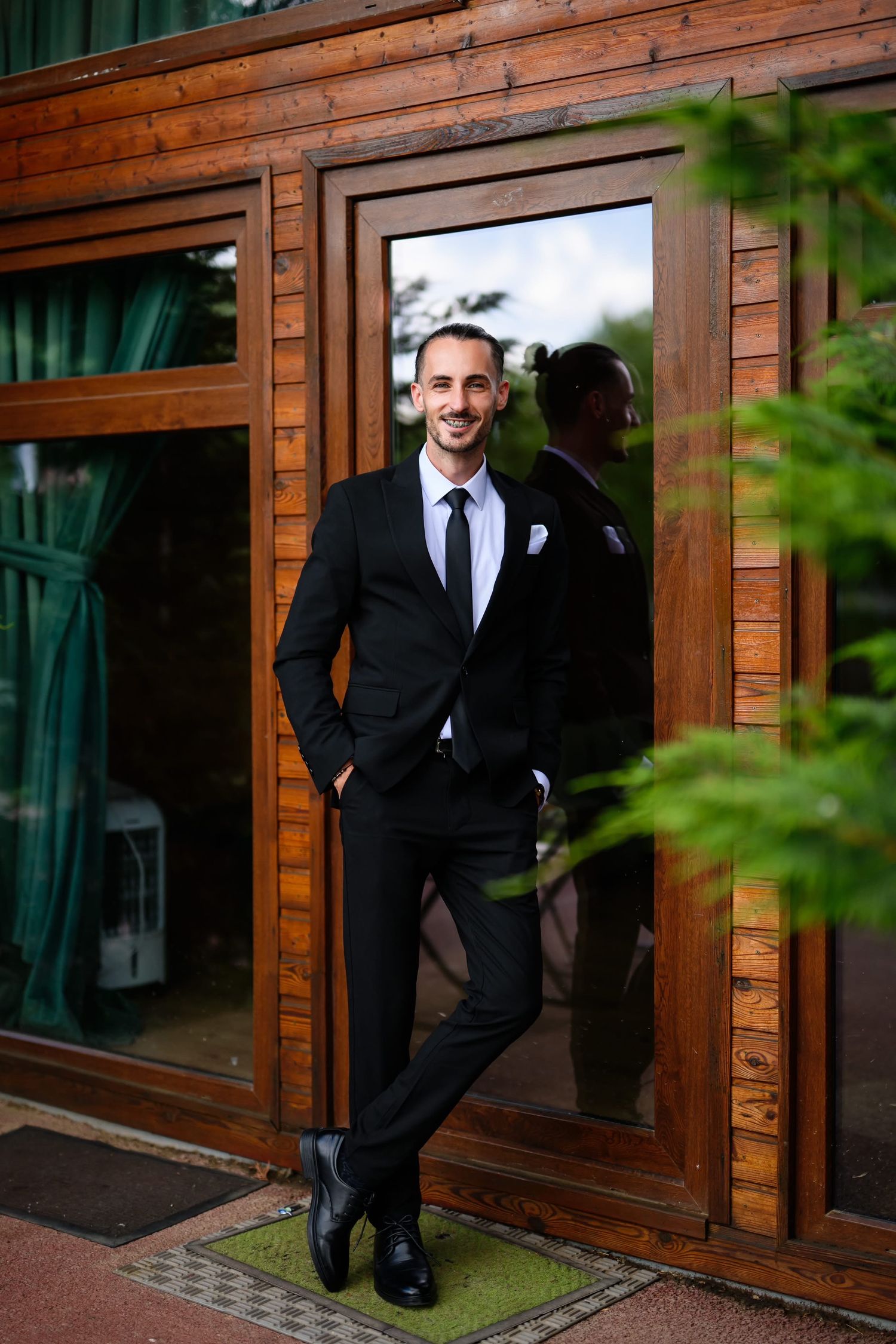 Smiling groom in black suit and tie posing confidently at wooden building entrance on wedding day.
