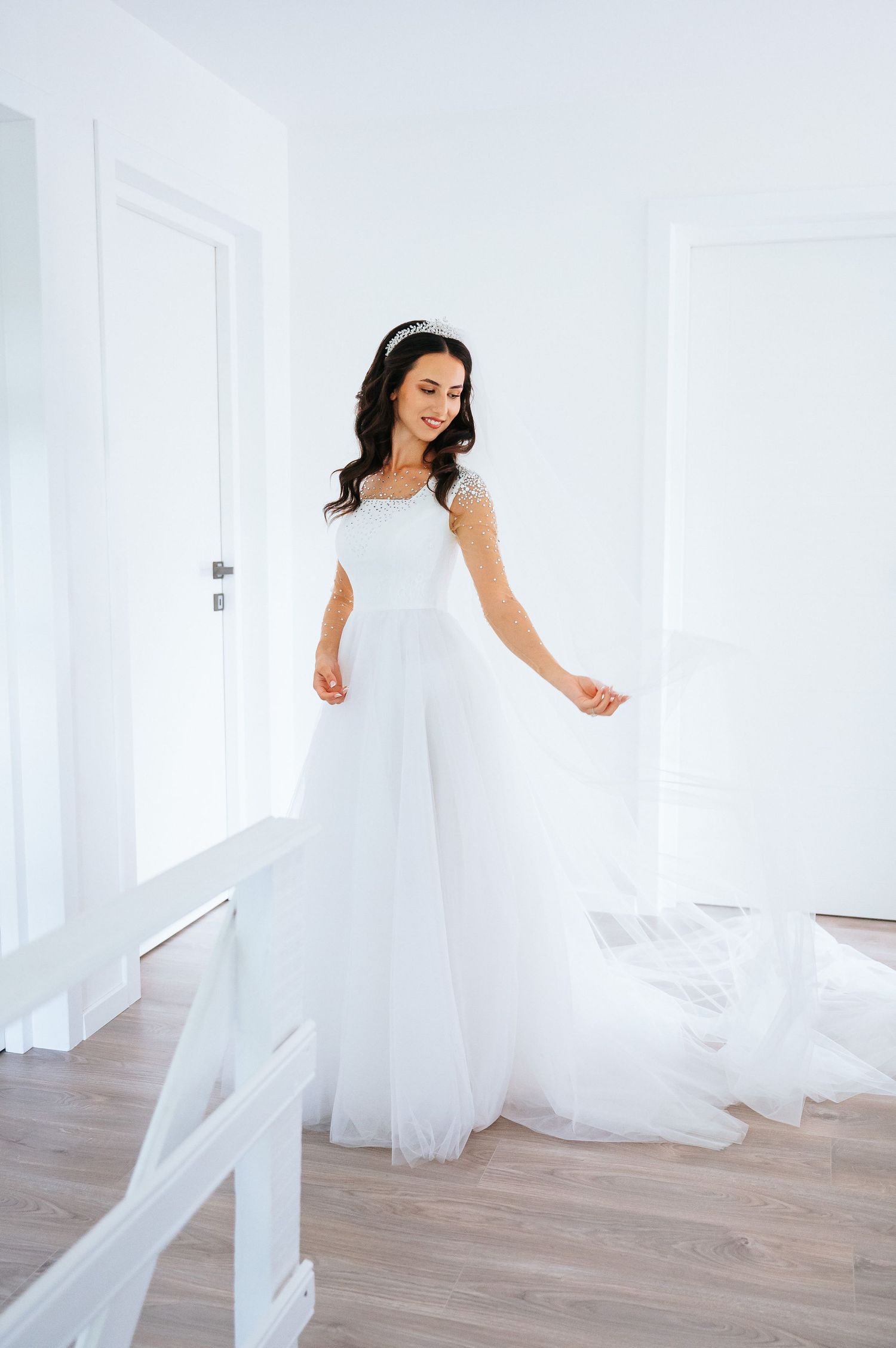 Elegant bride in a flowing white tulle wedding gown with a tiara, posing in a bright white room.