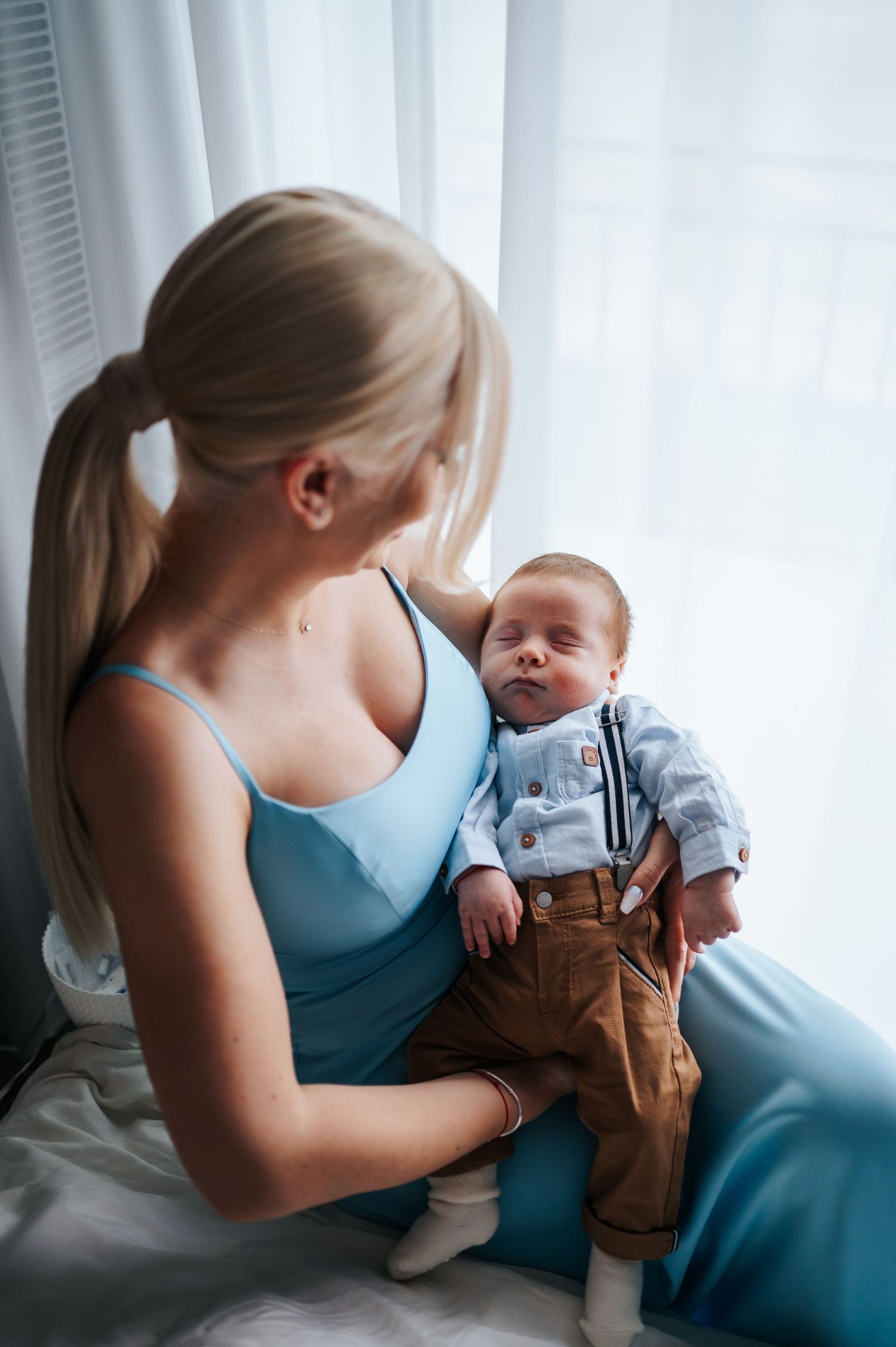 Blonde mother in blue dress tenderly holds newborn baby dressed in suspenders near a bright window.