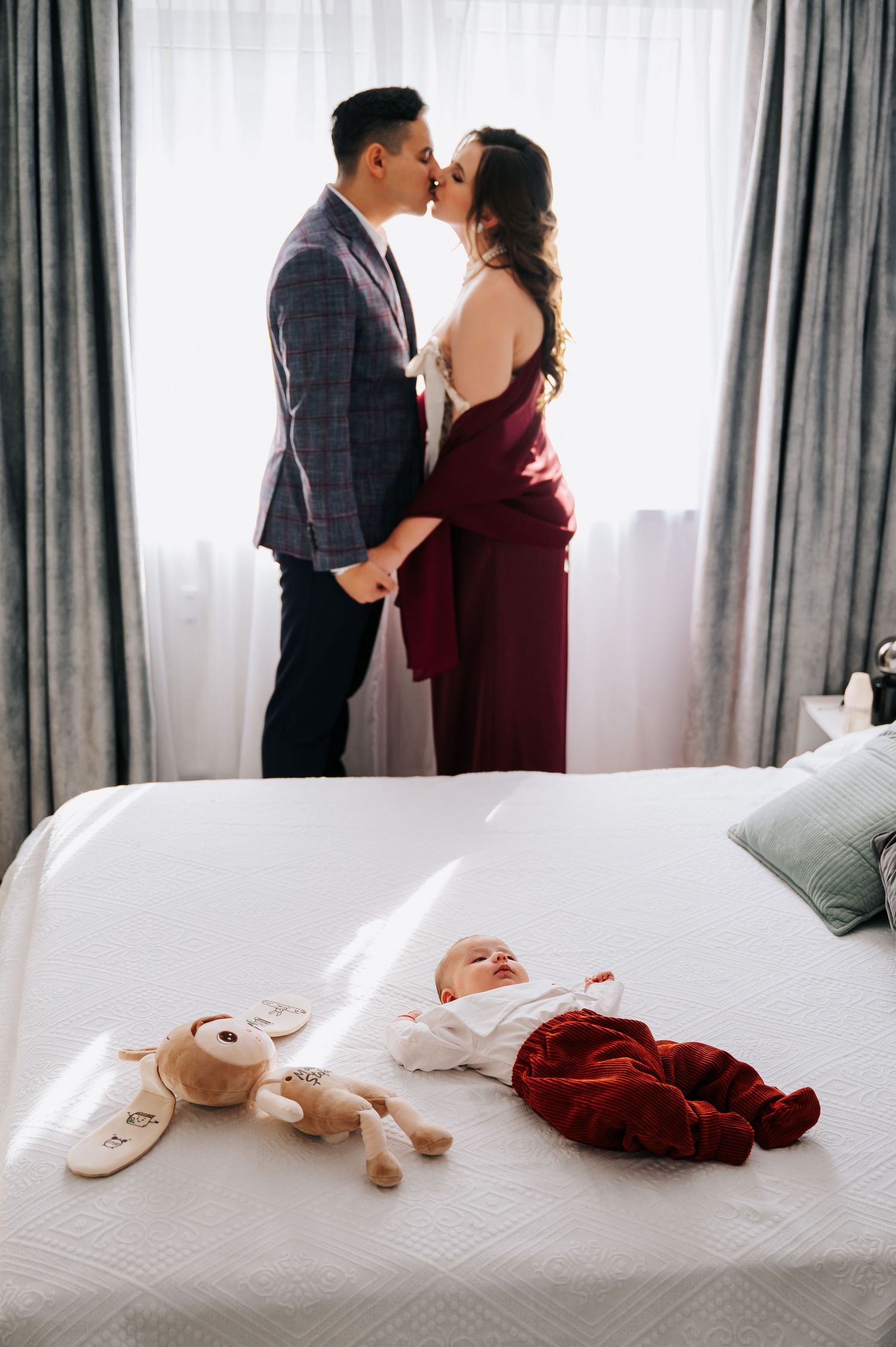 Couple kissing by window in bedroom while newborn baby lies on white bed with stuffed toy.
