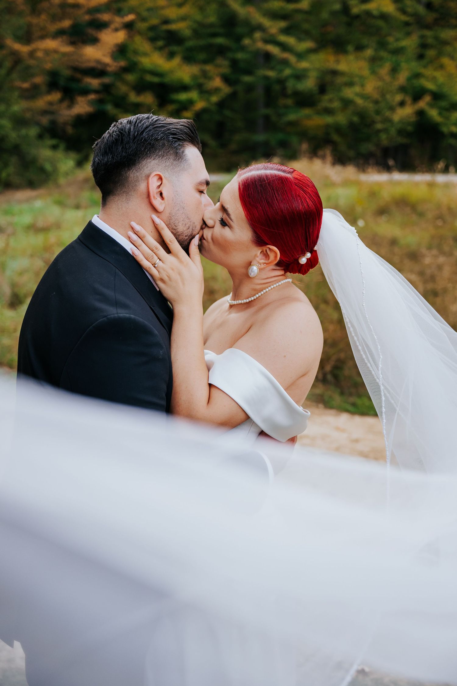 A bride with red hair and groom in black suit share a romantic kiss outdoors, with her white veil flowing in the breeze.