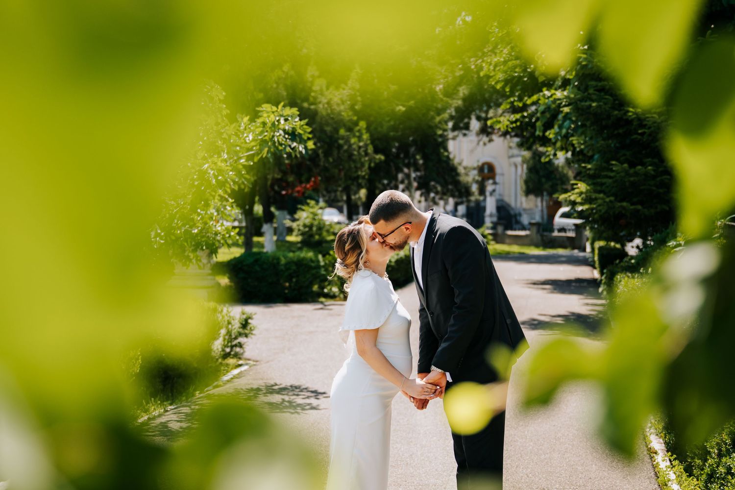 Romantic couple kissing on garden path framed by lush green leaves in soft sunlight.