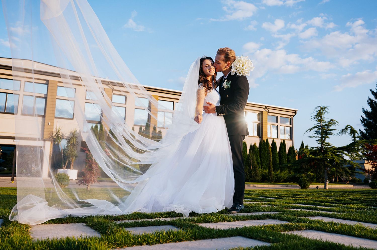 Bride and groom kissing with flowing veil in beautiful garden outside elegant wedding venue.