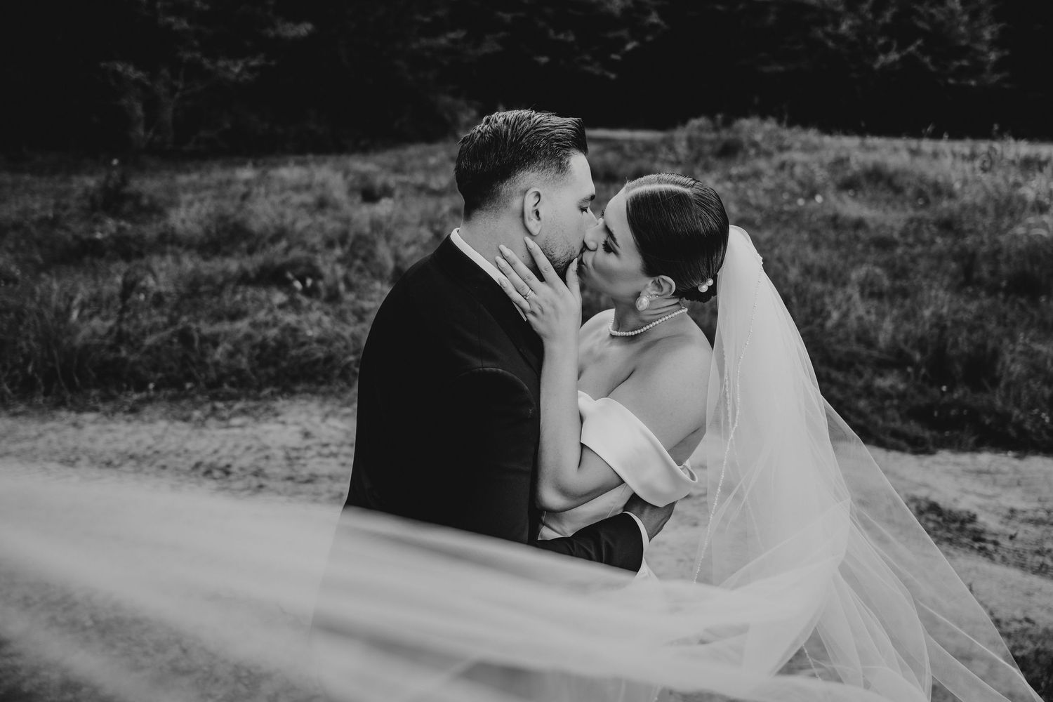 Bride and groom share romantic kiss outdoors as wedding veil blows in the wind.