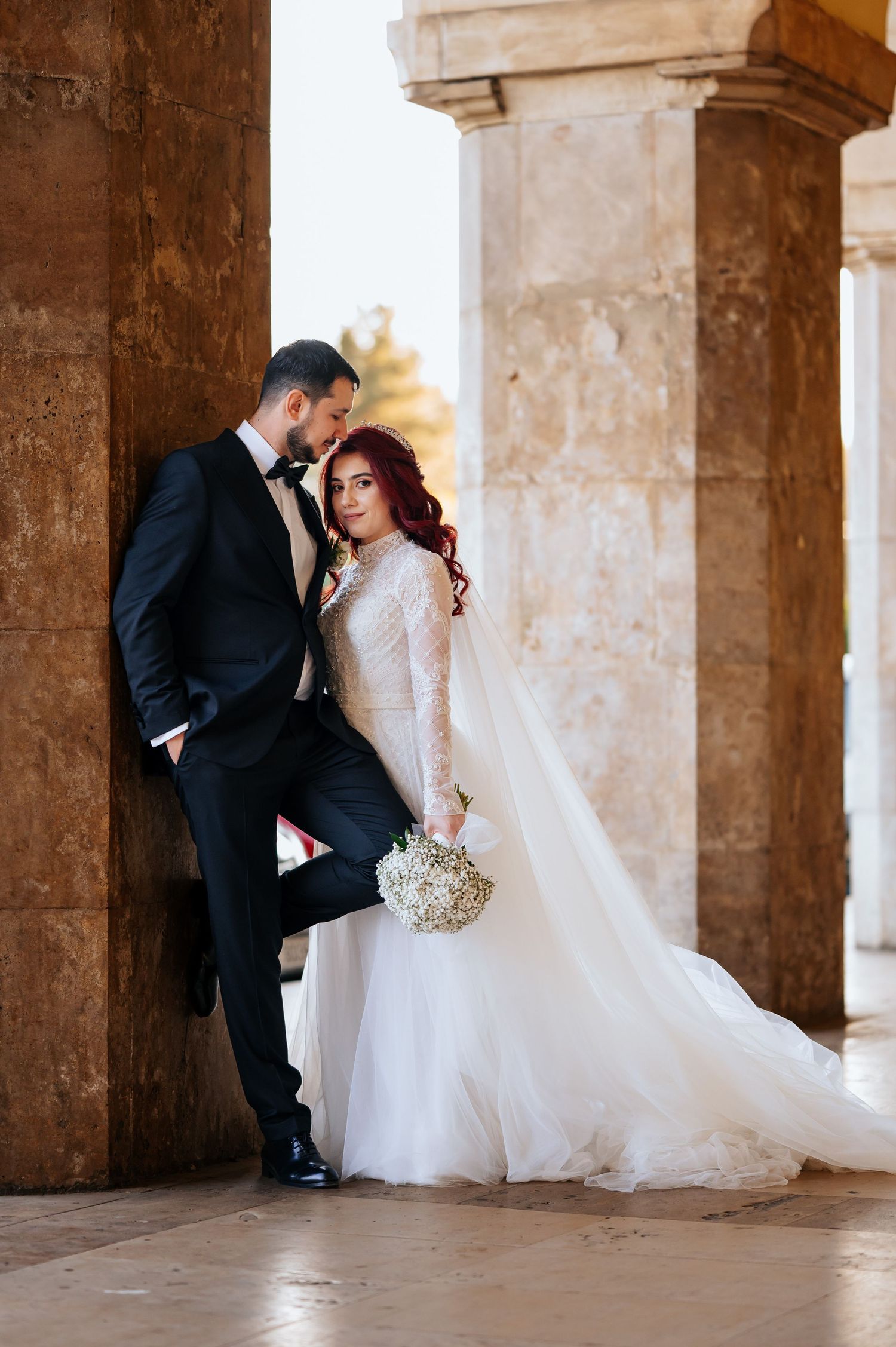 Bride in white gown with red hair holds bouquet beside groom in black tuxedo at stone column venue.