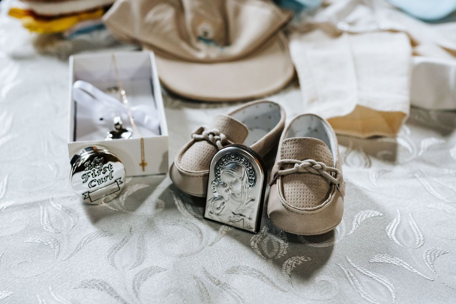Baby baptism shoes, silver religious icon, and cross necklace displayed on white lace tablecloth for christening ceremony.