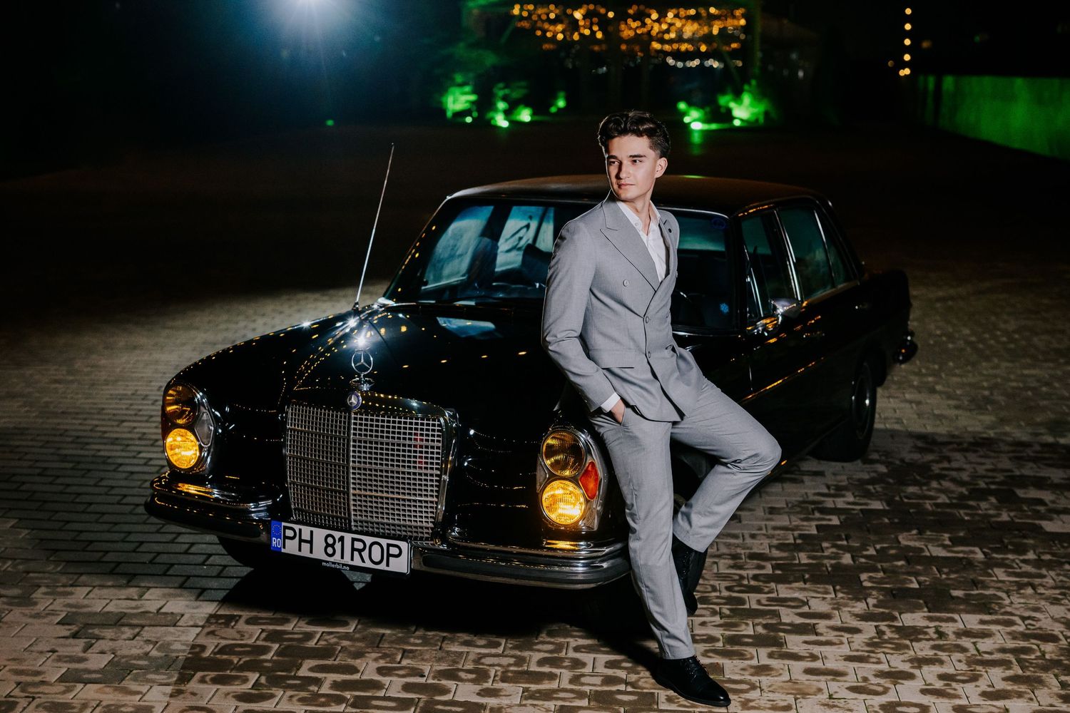 Young man in gray suit posing confidently against a classic black Mercedes-Benz at night.
