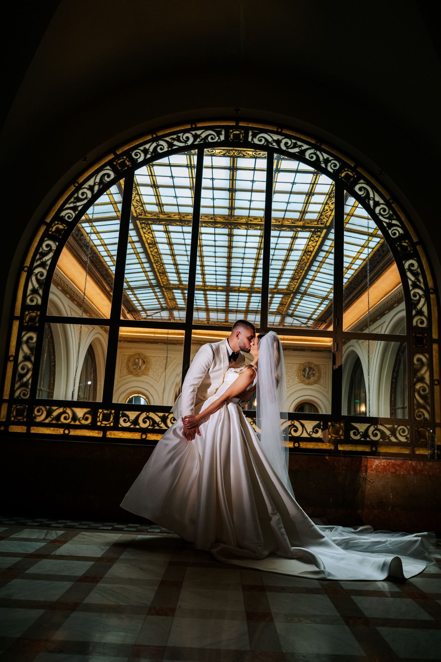 Bride and groom share a kiss beneath a stunning ornate stained glass arched window in a grand hall.