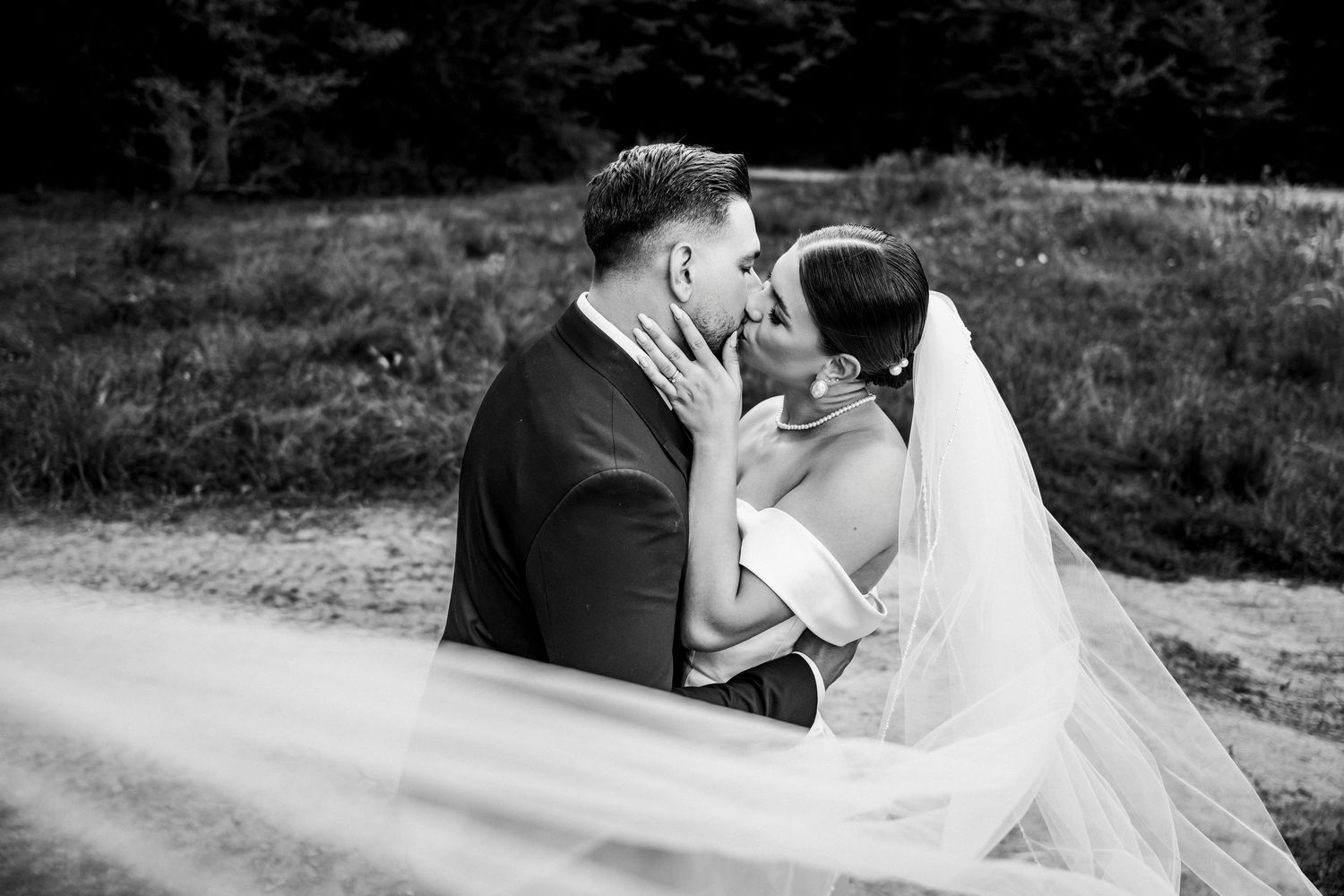 Black and white photo of a bride and groom sharing a romantic kiss outdoors, with her veil flowing in the breeze.