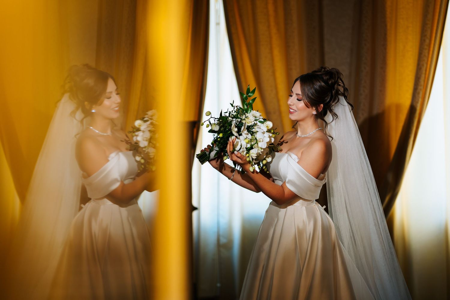 Radiant bride in a satin gown holds a bouquet while admiring her reflection in a gold-curtained mirror.
