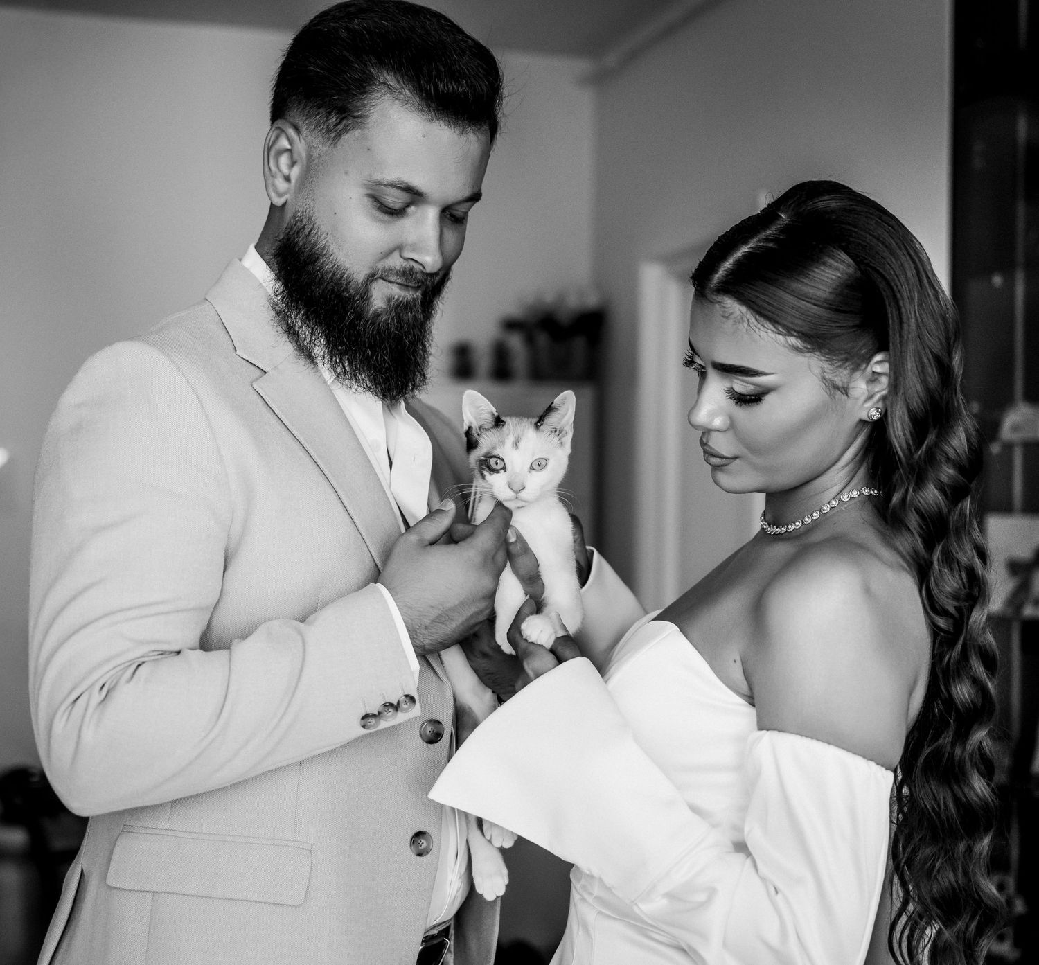Black and white photo of a bearded groom and bride in wedding attire holding a small kitten together.