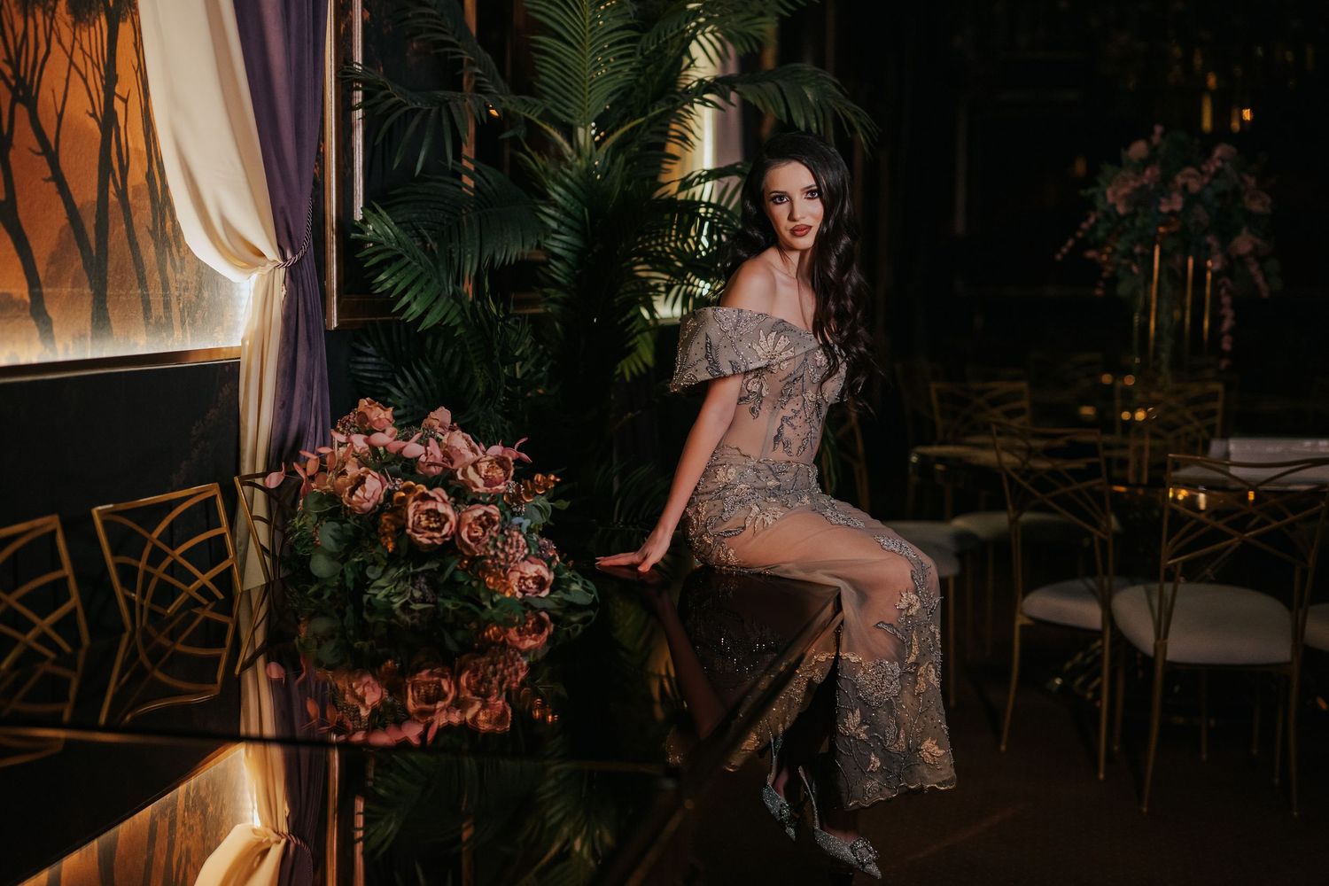 A woman in an elegant sheer floral gown sits gracefully beside pink roses in a luxurious dark botanical setting.