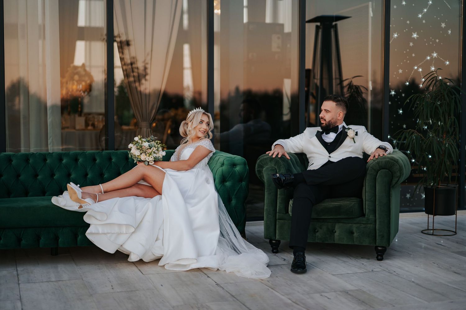Bride and groom in elegant wedding attire relaxing on green velvet furniture at a modern venue.