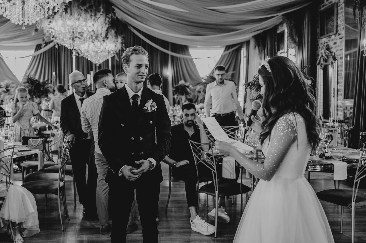 Black and white wedding photo of groom in naval uniform listening to bride read vows in elegant ballroom.