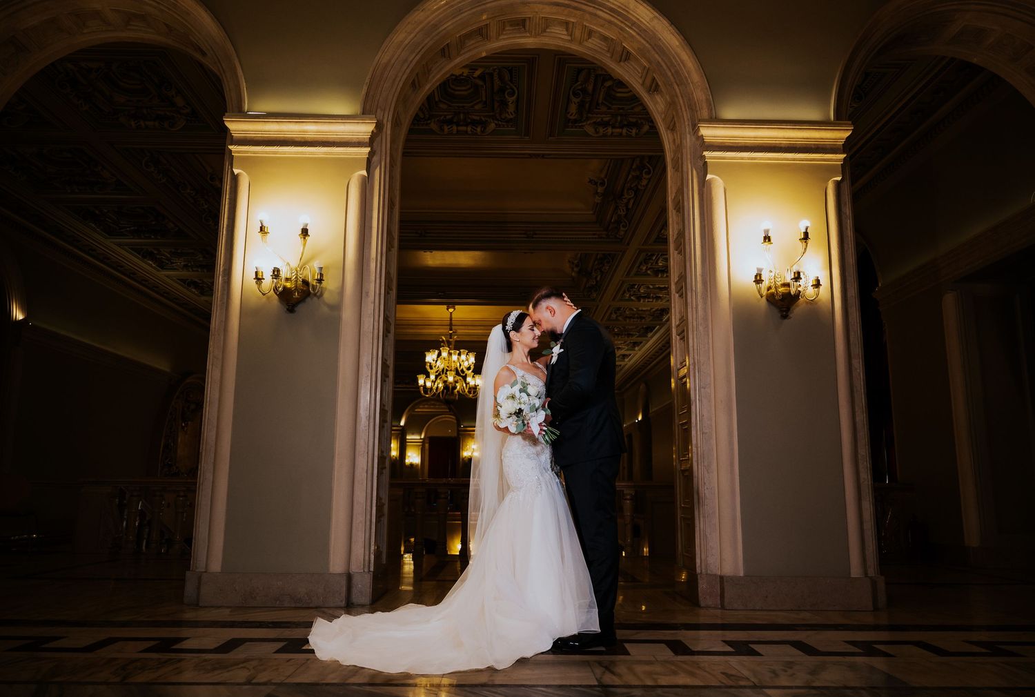 Bride and groom share a romantic kiss under golden arched hallway in an elegant wedding venue.